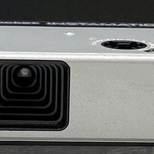 May include: A silver Kodak Pocket Instamatic 10 camera. The camera has a black lens, viewfinder, and a black shutter button. The Kodak logo is visible on the front. The camera is rectangular and has a retro design.