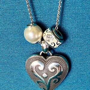 May include: A silver-toned necklace featuring a large heart-shaped pendant with a textured, filigree-style surface. The chain includes two decorative beads: one smooth, cream-colored spherical bead and one silver-toned bead with engraved patterns.