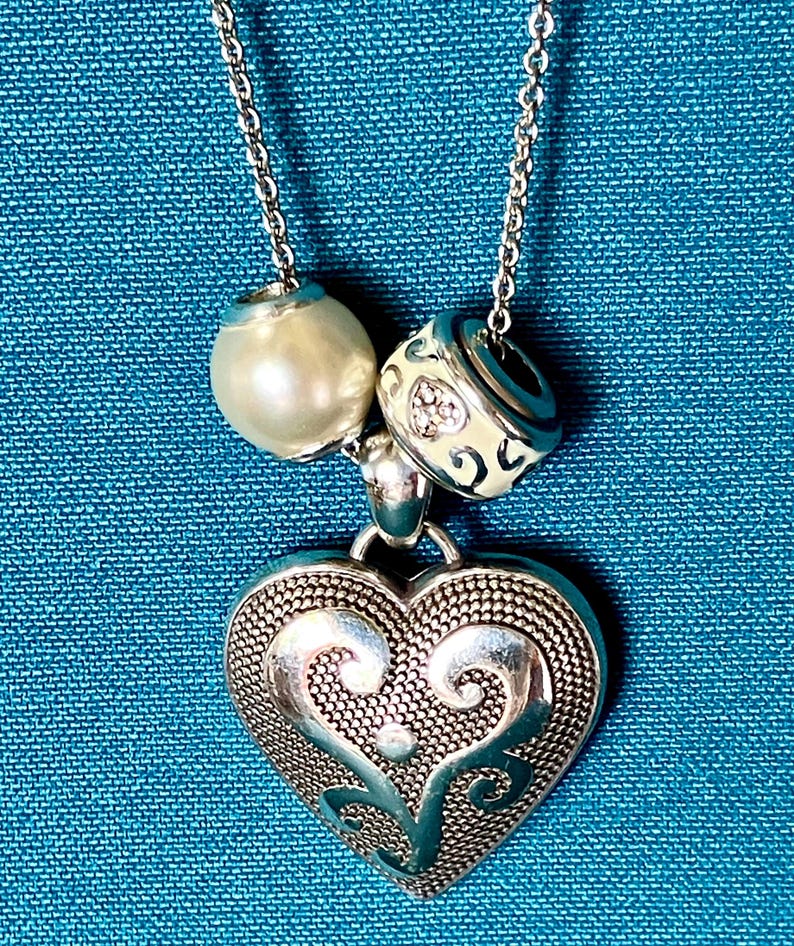 May include: A silver-toned necklace featuring a large heart-shaped pendant with a textured, filigree-style surface. The chain includes two decorative beads: one smooth, cream-colored spherical bead and one silver-toned bead with engraved patterns.