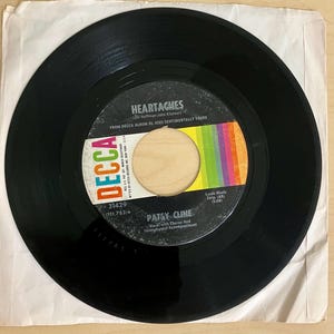 May include: A black vinyl record with the title "Heartaches" by Patsy Cline. The record label features the Decca logo, a colorful rainbow design, and the text "Vocal with Chorus And Instrumental Accompaniment."
