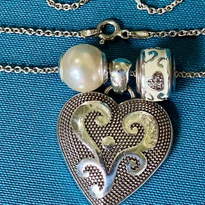 May include: A silver chain necklace featuring a large heart-shaped pendant with a textured background and smooth silver scrollwork. The necklace includes two beads: one round white pearl and one white barrel-shaped bead with small heart cutouts.