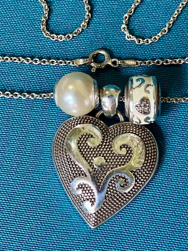 May include: A silver chain necklace featuring a large heart-shaped pendant with a textured background and smooth silver scrollwork. The necklace includes two beads: one round white pearl and one white barrel-shaped bead with small heart cutouts.