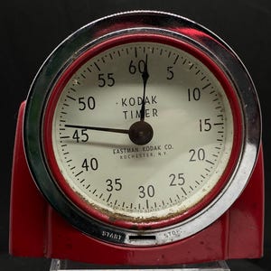 May include: A red and chrome vintage Kodak timer with a white face and black hands. The timer has a start and stop button and the text "Kodak Timer" and "Eastman Kodak Co. Rochester, N.Y." printed on the face.