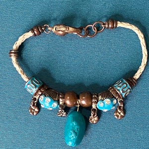 May include: A bracelet featuring a braided cream-colored cord, copper-toned metal beads, and turquoise-colored accents. The design includes two round brown beads, small dangling flower charms, and a central, irregular turquoise-colored stone pendant.