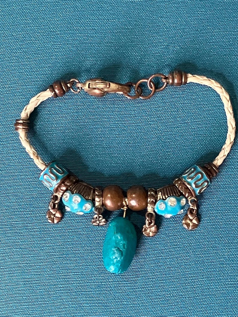 May include: A bracelet featuring a braided cream-colored cord, copper-toned metal beads, and turquoise-colored accents. The design includes two round brown beads, small dangling flower charms, and a central, irregular turquoise-colored stone pendant.