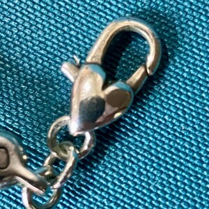 K&ouml;nnte beinhalten: Nahaufnahme eines silberfarbenen Verschlusses mit herzf&ouml;rmigem Design. Der Verschluss ist mit einer Kette verbunden, vor einem strukturierten t&uuml;rkisfarbenen Stoffhintergrund.