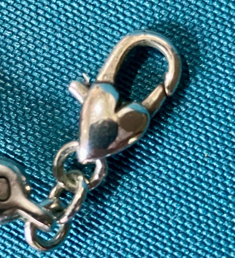 K&ouml;nnte beinhalten: Nahaufnahme eines silberfarbenen Verschlusses mit herzf&ouml;rmigem Design. Der Verschluss ist mit einer Kette verbunden, vor einem strukturierten t&uuml;rkisfarbenen Stoffhintergrund.
