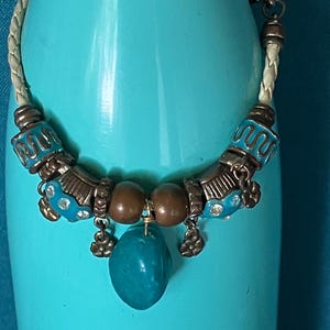May include: A bracelet featuring a braided cream-colored cord, accented with metallic bronze-toned beads, turquoise-colored enamel spacers with wavy patterns, small flower-shaped charms, and a central smooth, irregular turquoise-colored stone pendant.