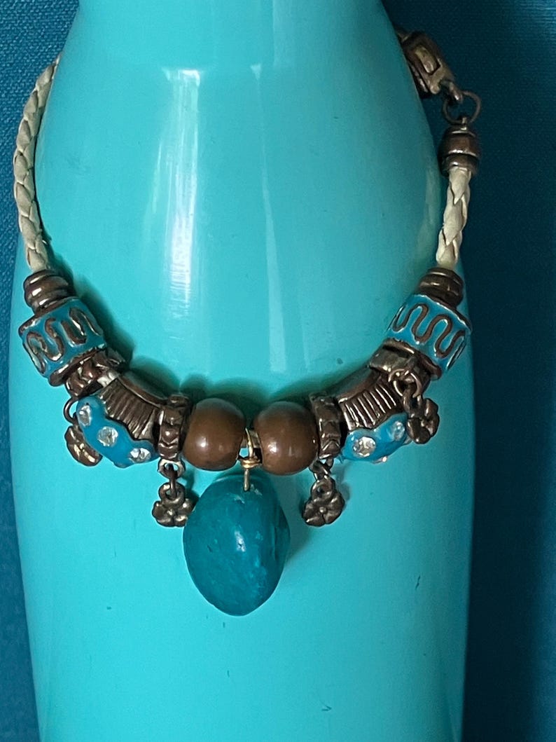 May include: A bracelet featuring a braided cream-colored cord, accented with metallic bronze-toned beads, turquoise-colored enamel spacers with wavy patterns, small flower-shaped charms, and a central smooth, irregular turquoise-colored stone pendant.
