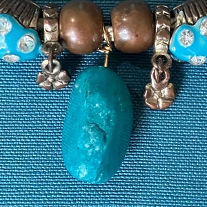 May include: A bracelet featuring a central rough-cut turquoise stone pendant, flanked by two smooth copper-toned beads, two silver-toned textured spacers, two small flower-shaped charms, and two bright blue beads embedded with clear crystals.