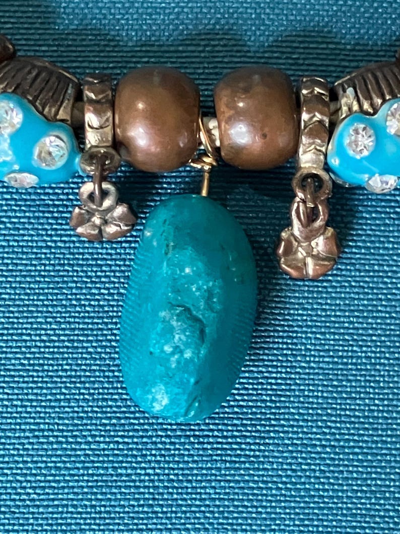 May include: A bracelet featuring a central rough-cut turquoise stone pendant, flanked by two smooth copper-toned beads, two silver-toned textured spacers, two small flower-shaped charms, and two bright blue beads embedded with clear crystals.