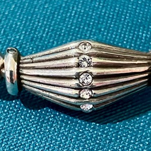 K&ouml;nnte beinhalten: Nahaufnahme eines silbernen Armbands. Die zentrale, geriffelte Perle ist mit f&uuml;nf kleinen, klaren Kristallen besetzt. Das Armband liegt auf einem t&uuml;rkisfarbenen Hintergrund, der den Glanz des Silbers und das Funkeln der Kristalle hervorhebt.