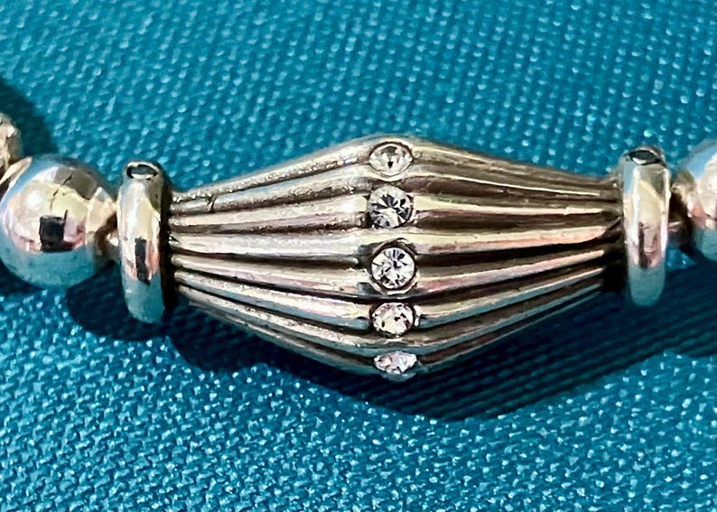 K&ouml;nnte beinhalten: Nahaufnahme eines silbernen Armbands. Die zentrale, geriffelte Perle ist mit f&uuml;nf kleinen, klaren Kristallen besetzt. Das Armband liegt auf einem t&uuml;rkisfarbenen Hintergrund, der den Glanz des Silbers und das Funkeln der Kristalle hervorhebt.