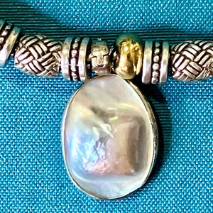 May include: A necklace featuring a central oval-shaped iridescent white pendant suspended from a strand of alternating silver-toned metal beads. The beads include textured woven patterns and smooth metallic spacers, creating a detailed design.