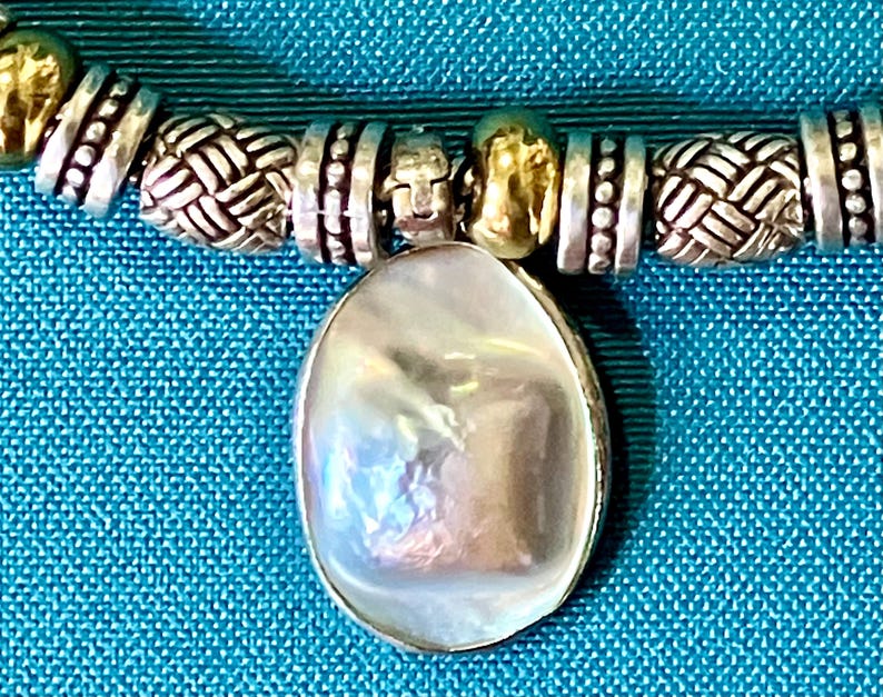 May include: A necklace featuring a central oval-shaped iridescent white pendant suspended from a strand of alternating silver-toned metal beads. The beads include textured woven patterns and smooth metallic spacers, creating a detailed design.