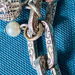 May include: A silver-toned chain necklace featuring rectangular links with an embossed scrollwork pattern. The chain is adorned with a small, round, white pearl-like bead and a spherical, open-work metal charm attached to the links by jump rings.
