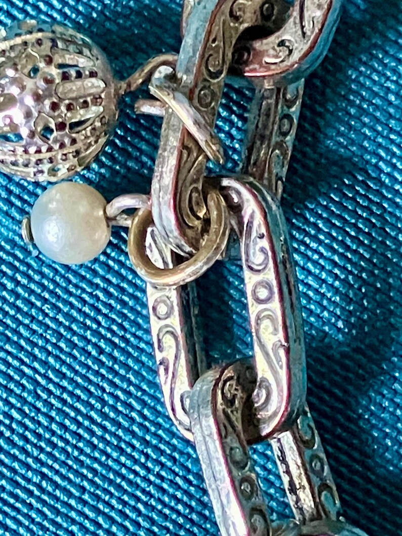 May include: A silver-toned chain necklace featuring rectangular links with an embossed scrollwork pattern. The chain is adorned with a small, round, white pearl-like bead and a spherical, open-work metal charm attached to the links by jump rings.