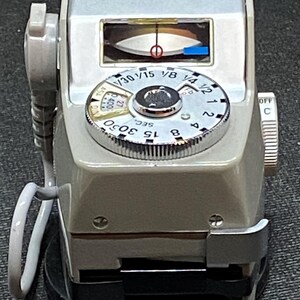 May include: A Canon Booster light meter with a white body and a silver dial. The dial has markings for shutter speeds and aperture settings. The meter has a needle that points to the correct exposure setting.
