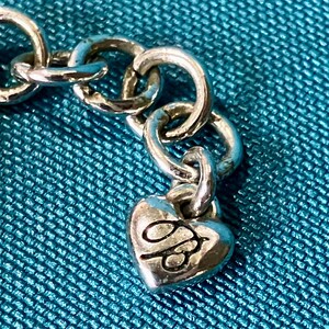 K&ouml;nnte beinhalten: Nahaufnahme einer silberfarbenen Kette mit einem herzf&ouml;rmigen Anh&auml;nger. Der Anh&auml;nger hat ein gepr&auml;gtes Design und ist mit einem kleinen Ring an der Kette befestigt. Der Hintergrund ist ein strukturierter t&uuml;rkisfarbener Stoff.