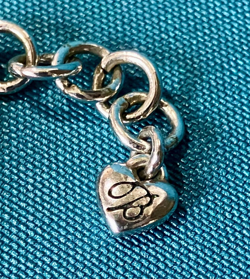K&ouml;nnte beinhalten: Nahaufnahme einer silberfarbenen Kette mit einem herzf&ouml;rmigen Anh&auml;nger. Der Anh&auml;nger hat ein gepr&auml;gtes Design und ist mit einem kleinen Ring an der Kette befestigt. Der Hintergrund ist ein strukturierter t&uuml;rkisfarbener Stoff.