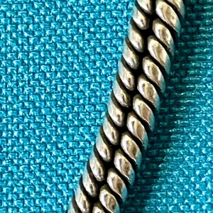 May include: A silver-toned metal chain featuring a dense, braided rope-like texture with a polished finish. The chain consists of tightly interlocking links that create a continuous, rounded, and flexible cylindrical form with deep grooves.