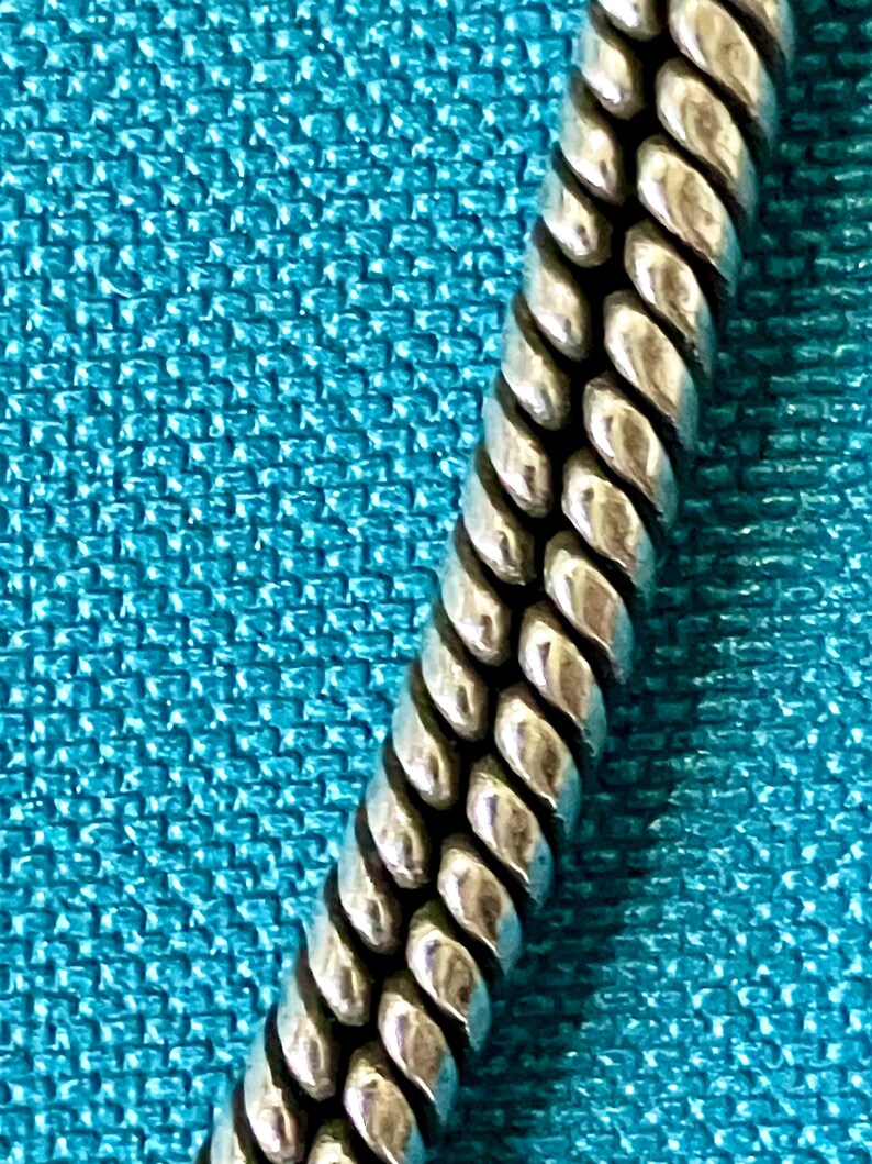 May include: A silver-toned metal chain featuring a dense, braided rope-like texture with a polished finish. The chain consists of tightly interlocking links that create a continuous, rounded, and flexible cylindrical form with deep grooves.