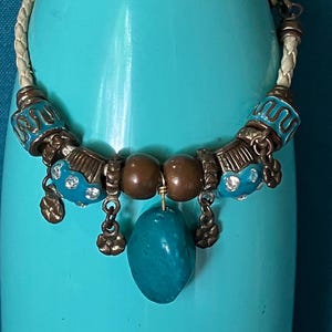 May include: A bracelet featuring a braided cream-colored cord, accented with metallic bronze-toned beads, turquoise-colored enamel beads with embedded clear crystals, and a central smooth, irregular turquoise-colored stone pendant.