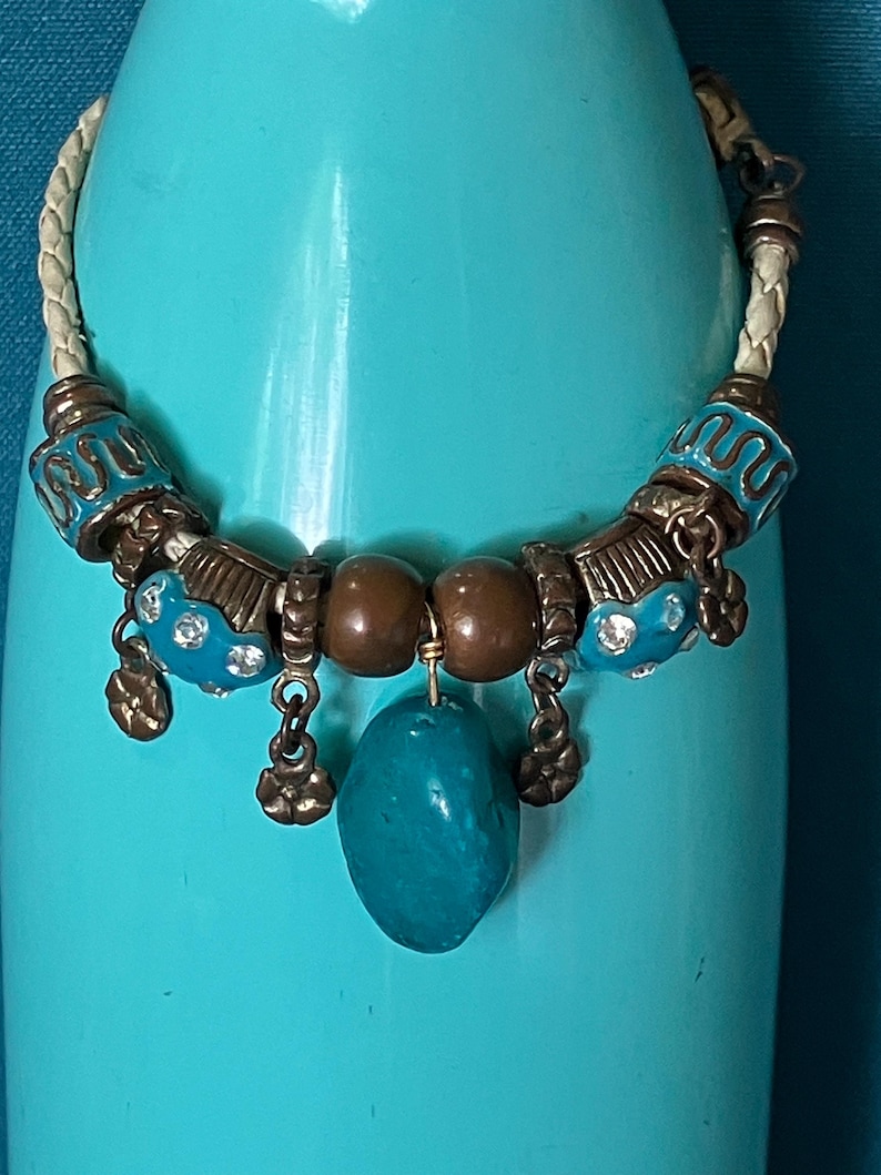 May include: A bracelet featuring a braided cream-colored cord, accented with metallic bronze-toned beads, turquoise-colored enamel beads with embedded clear crystals, and a central smooth, irregular turquoise-colored stone pendant.