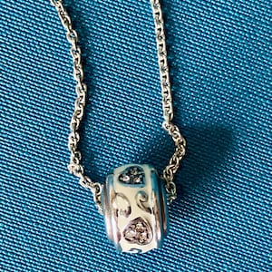 May include: A silver-toned metal chain necklace featuring a cylindrical bead charm. The bead has a white enamel finish with silver scrollwork patterns and two heart-shaped insets filled with small, clear, sparkling stones.
