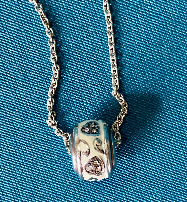 May include: A silver-toned metal chain necklace featuring a cylindrical bead charm. The bead has a white enamel finish with silver scrollwork patterns and two heart-shaped insets filled with small, clear, sparkling stones.