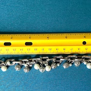 May include: A silver-toned chain link bracelet featuring dangling white pearl beads and ornate silver spheres. The bracelet is positioned below a yellow ruler, showing a total length of approximately 19 cm from end to end.