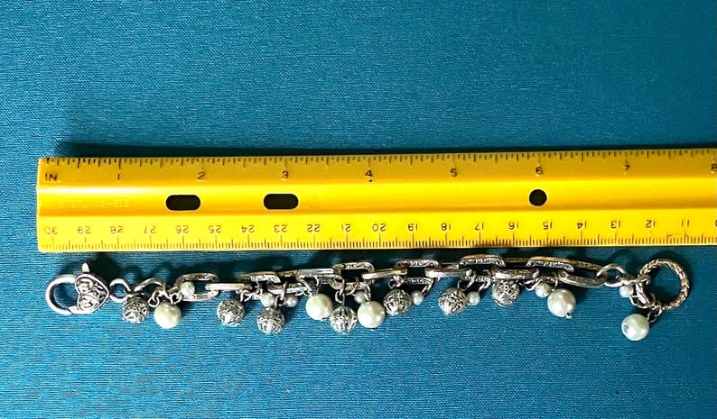 May include: A silver-toned chain link bracelet featuring dangling white pearl beads and ornate silver spheres. The bracelet is positioned below a yellow ruler, showing a total length of approximately 19 cm from end to end.