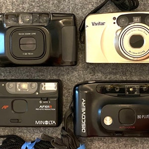 May include: Four different film cameras, a black Ricoh, a silver Vivitar, a black Minolta, and a black Fuji. The Vivitar camera has the text "530 PZ DATE A PRINT" on it. The Fuji camera has the text "90 PLUS AUTO FOCUS" on it.