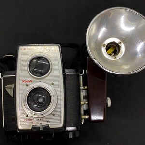 May include: Vintage Kodak Brownie Reflex camera with a flash attachment. The camera has a silver face with two lenses and a black body. The flash is silver with a dark brown base.