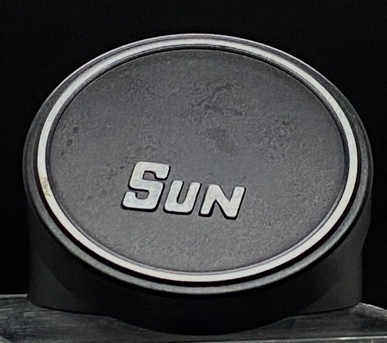 Puede incluir: Un altavoz redondo negro con un borde plateado y la palabra "SUN" impresa en letras plateadas.