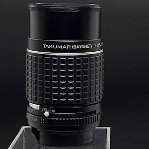 May include: A black camera lens with silver accents. The lens is labeled "Takumar (Bayonet) 1:2.5 135 mm".