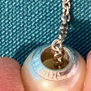 May include: A spherical white pearl bead with a silver metal core featuring the engraved text 925, suspended from a delicate silver chain. The bead has a smooth, lustrous surface and a central opening through which the metal chain passes.