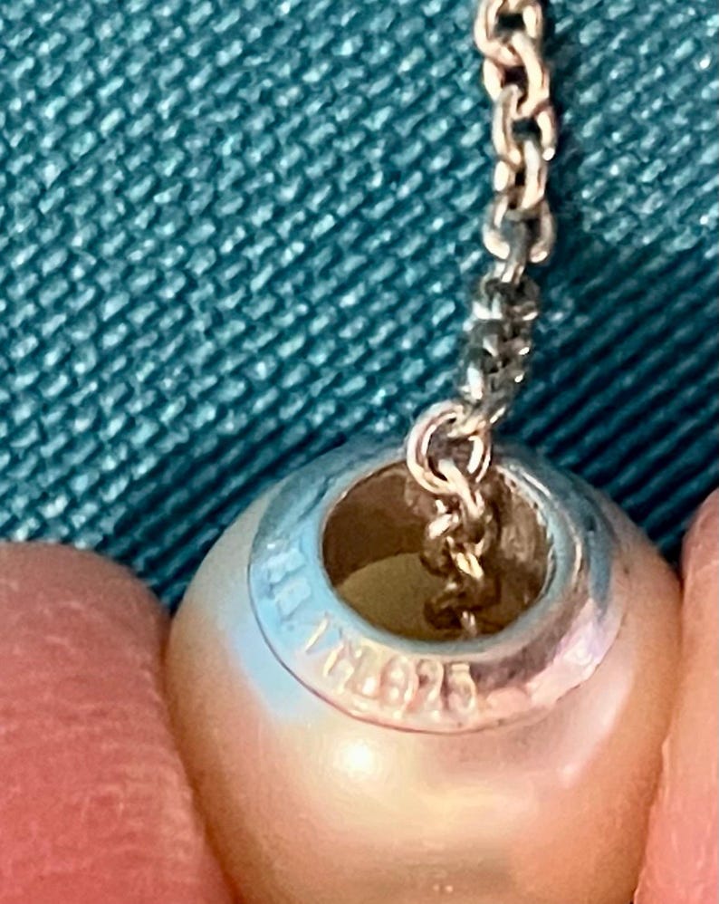 May include: A spherical white pearl bead with a silver metal core featuring the engraved text 925, suspended from a delicate silver chain. The bead has a smooth, lustrous surface and a central opening through which the metal chain passes.
