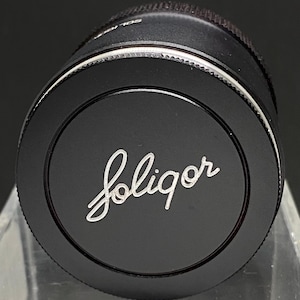 May include: A black camera lens with a silver ring around the edge. The lens has the word "Soligor" written in white on the front.
