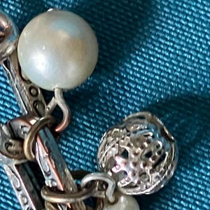 May include: A silver-toned charm bracelet featuring a series of interconnected links with an intricate embossed pattern. The bracelet is adorned with several round, white, lustrous beads and a spherical, open-work silver charm with a textured surface.