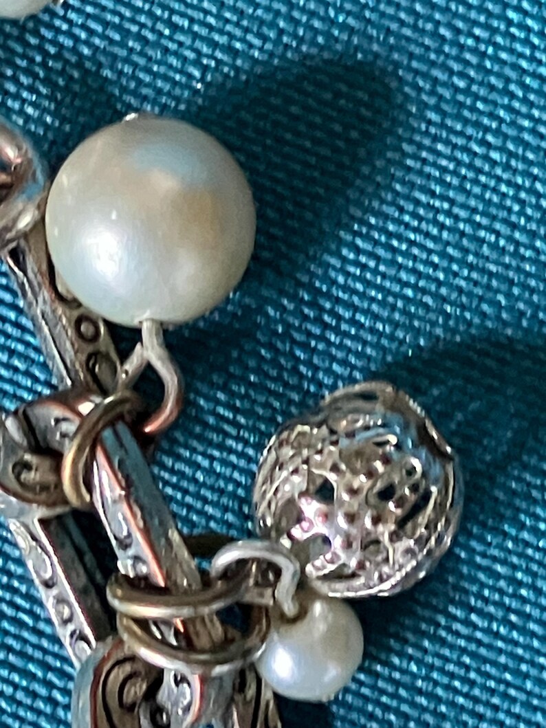 May include: A silver-toned charm bracelet featuring a series of interconnected links with an intricate embossed pattern. The bracelet is adorned with several round, white, lustrous beads and a spherical, open-work silver charm with a textured surface.