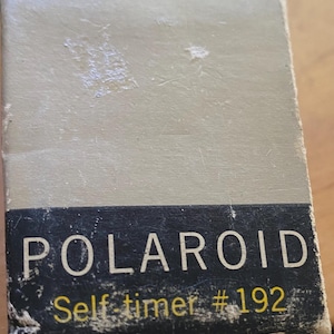May include: A vintage Polaroid self-timer box. The box is rectangular with a faded beige color and a black label with the word "POLAROID" in white letters. Below, in yellow, is "Self-timer #192".