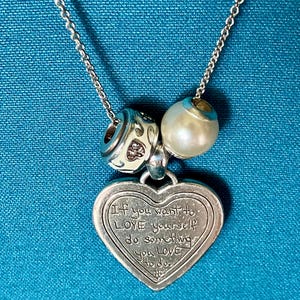 May include: A silver-toned necklace featuring a heart-shaped pendant engraved with the text 'If you want to LOVE yourself do something you LOVE to do.' The chain includes two charms: one white bead with a metallic pattern and one smooth white pearl-like bead.
