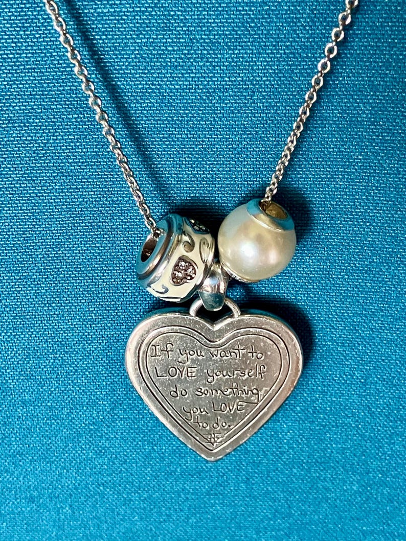 May include: A silver-toned necklace featuring a heart-shaped pendant engraved with the text 'If you want to LOVE yourself do something you LOVE to do.' The chain includes two charms: one white bead with a metallic pattern and one smooth white pearl-like bead.