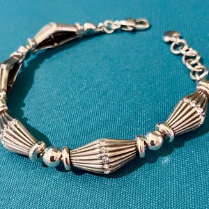 K&ouml;nnte beinhalten: Silbernes Armband mit einer Reihe von dekorativen, f&auml;cherf&ouml;rmigen Gliedern, die mit runden Perlen und kleinen, klaren Edelsteinen durchsetzt sind. Das Armband hat einen Karabinerverschluss und eine verstellbare Kette.