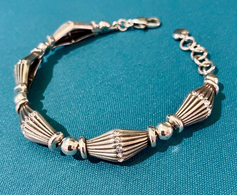 K&ouml;nnte beinhalten: Silbernes Armband mit einer Reihe von dekorativen, f&auml;cherf&ouml;rmigen Gliedern, die mit runden Perlen und kleinen, klaren Edelsteinen durchsetzt sind. Das Armband hat einen Karabinerverschluss und eine verstellbare Kette.