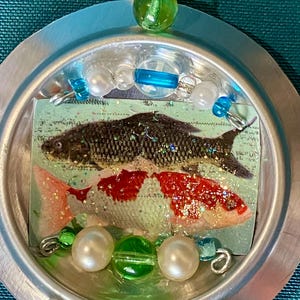 May include: A handmade pendant featuring a silver-colored metal frame. Inside, a picture of two koi fish is visible. The pendant is adorned with green and white beads, and small blue accents. The pendant hangs from a white bead.