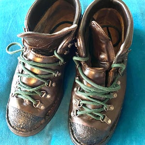 May include: A pair of dark brown leather boots featuring green laces, metal eyelets, and black textured rubber toe caps. The boots have a high-top design with padded collars and visible stitching along the base of the soles.