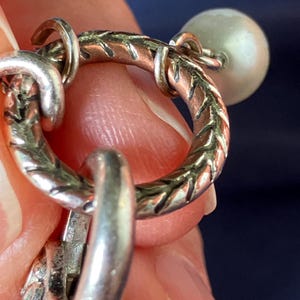 May include: A circular silver-toned metal ring featuring a textured, etched pattern along its surface. The ring is connected to a small, smooth, off-white spherical bead and a larger, ornate, textured silver-toned metal component hanging below it.