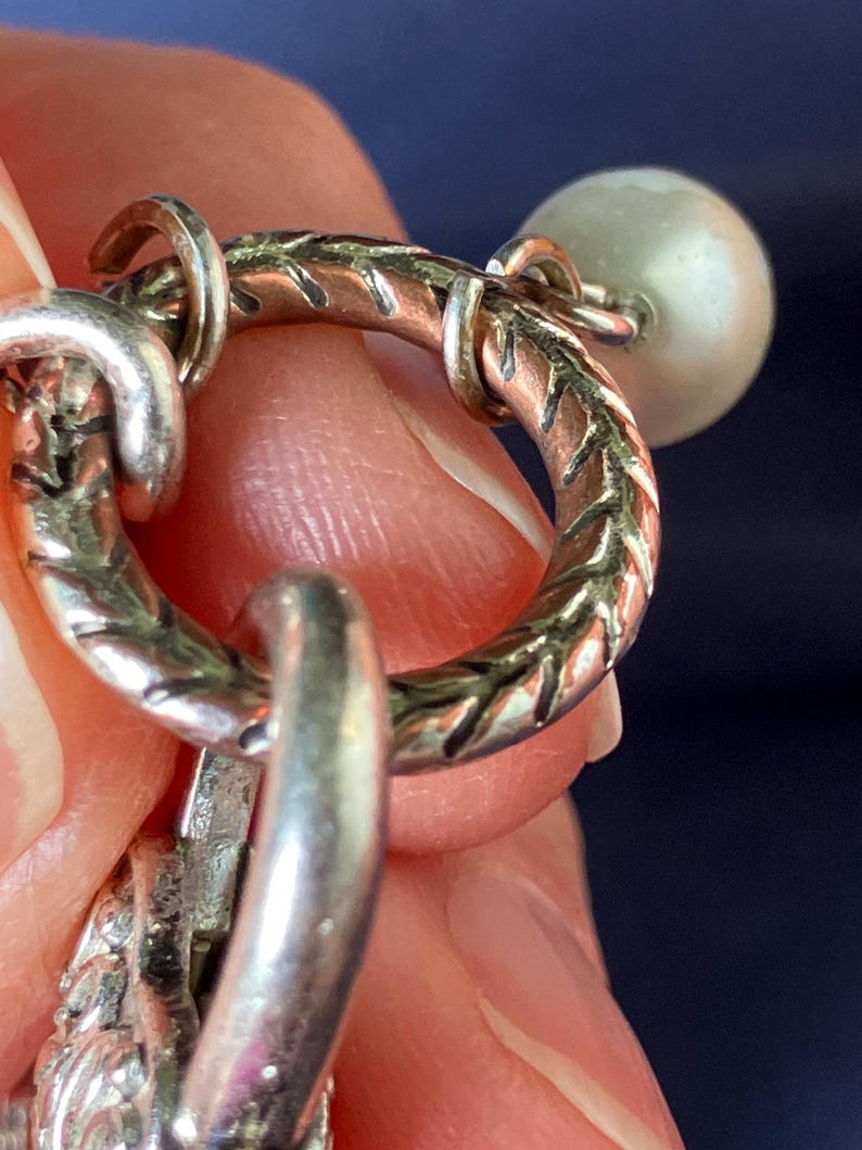 May include: A circular silver-toned metal ring featuring a textured, etched pattern along its surface. The ring is connected to a small, smooth, off-white spherical bead and a larger, ornate, textured silver-toned metal component hanging below it.